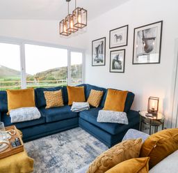 A living room with a blue corner sofa and mustard cushions by large windows at Pennant Cottage in Llithfaen