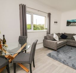 A living room with a glass dining table and grey chairs next to a grey corner sofa and a blue rug at Ty Cariad in Llangynin near St Clears