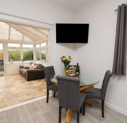A dining area with a glass table and four chairs with a TV mounted on the wall and a conservatory with a sofa and glass door at Ty Cariad in Llangynin near St Clears
