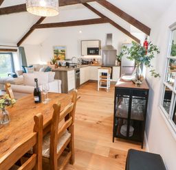 An open plan kitchen and dining area with wooden dining table and chairs a kitchen island white cabinets sofa and large window at Carines Barns - Pol Cubert
