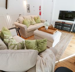 A living room with two sofas, green cushions, an ottoman with a wooden tray, a wall mounted TV and a small stove at Carines Barns - Par in Cubert