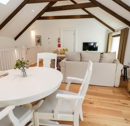 A dining area with a round white table and chairs and a living area with beige sofas and a wall-mounted TV in a room with wooden ceiling beams at Carines Barns - Par in Cubert