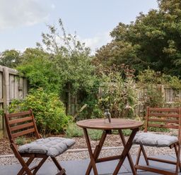 A small outdoor patio with two wooden chairs with cushions and a round wooden table with a glass vase surrounded by plants and a wooden fence at Carines Barns - Wesley Barn Cubert
