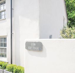 The exterior corner of a white cottage with windows plants and a sign reading Cob Cottage near Rock