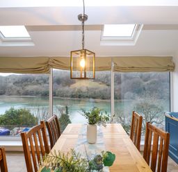 A dining room with a wooden table and chairs beside large windows overlooking a river view at Anjoma in Looe
