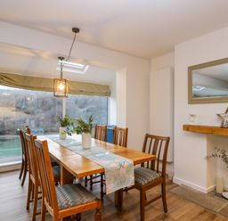 A dining room with wooden table and chairs by large windows overlooking a river at Anjoma in Looe