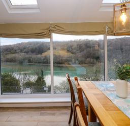 A dining area with wooden table and chairs by large windows overlooking a river and wooded hills at Anjoma in Looe