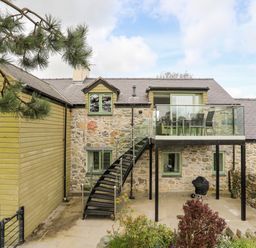 A stone and wooden house with an outdoor staircase leading to a glass balcony with a table and chairs at Gwalia Cottage in Brynsiencyn
