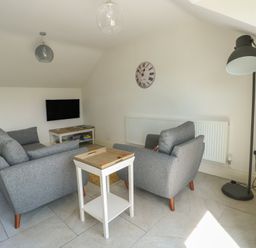 A living room with grey sofas a small white side table a flat screen tv on a white stand a floor lamp and a clock on the wall at Gwalia Cottage in Brynsiencyn