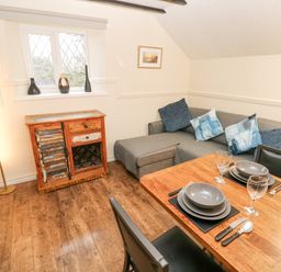 A dining area with a wooden table set for two next to a gray sofa with cushions and a wooden cabinet under a window at The Hayloft in St Florence