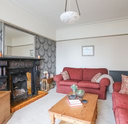 A living room with red sofas a wooden coffee table a fireplace with a large mirror above and patterned wallpaper at Llwyn Onn in Criccieth