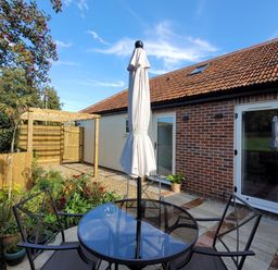 An outdoor patio with a glass table and four chairs with a closed umbrella in a garden at Beck View in Weston Longville near Lenwade