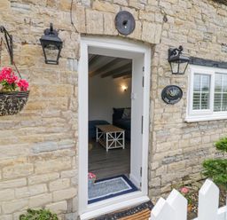 An open door with a view of a living room through a stone wall exterior with hanging flower basket and two black lanterns at The Cottage in Ettington