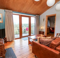 A living room with a brown sofa a wooden coffee table a television and glass doors opening to a balcony at Y Caban Glas in Rhydlewis near Brynhoffnant