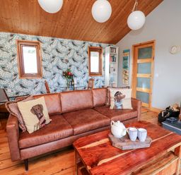 A living room with a brown sofa with dog print cushions a wooden coffee table with a teapot and cups and a wood stove at Y Caban Glas in Rhydlewis near Brynhoffnant