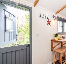 A small room with a blue stable door a wooden table and stools a window and coat hooks at Godrevy Shepherds Hut Angarrack near Hayle