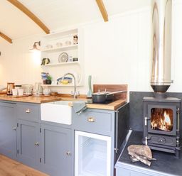 A kitchen area with blue cabinets a sink a stove and a wood burning stove with fire at Godrevy Shepherds Hut Angarrack near Hayle