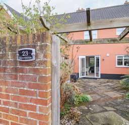 A brick wall with a house number plaque and a patio with wooden pergola and an open door at 23 Church Farm Lane in Halesworth