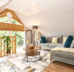 A living room with a gray sofa blue and yellow pillows a round wooden coffee table and a large window with orange patterned blinds at Strawberry Fields Annex in Emneth