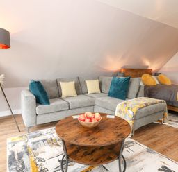 A living area with a grey sectional sofa and a wooden round coffee table with apples a bed with grey bedding and yellow pillows in the background at Strawberry Fields Annex in Emneth