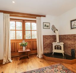 A room with a wooden floor a small stove in a brick corner a table with flowers and wooden doors at Oak Barn in Kentisbeare