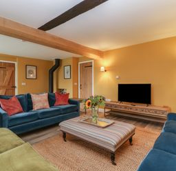 A living room with blue and green sofas a striped ottoman a wooden tv stand with a tv and a vase of flowers on the ottoman at Byways in Madresfield near Malvern