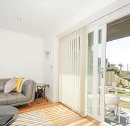 A living room with a grey sofa and pillows next to sliding glass doors opening to a patio with a table and chairs at Pebbles in Downderry