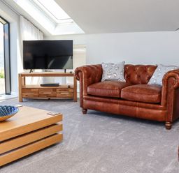 A living room with a brown leather sofa, a wooden coffee table, a television on a wooden stand, and a large window showing a sea view at The Waves in Amble