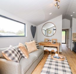 A living room with a sofa and cushions a wooden dining table with chairs a large round mirror and a kitchen area at Fawn's Rest in Nantmel near Rhayader
