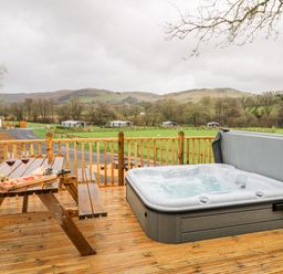 A wooden deck with a picnic table holding wine glasses and snacks next to a jacuzzi overlooking green fields and hills at Doe Lodge in Nantmel near Rhayader