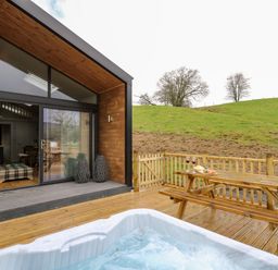 A wooden deck with a hot tub and picnic table next to a modern cabin and grassy hill at Doe Lodge in Nantmel near Rhayader