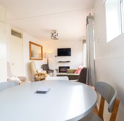 A dining area with chairs and round table leading to a living room with armchairs a sofa a wall mirror and a mounted tv at Sandpiper in Perranporth