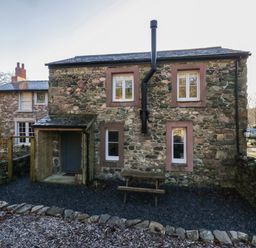 Exterior view of a stone cottage with four windows a wooden door under a small porch and a picnic table in a gravel yard at Church How Cottage in Nether Wasdale