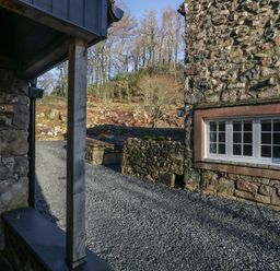 A gravel driveway between stone buildings with a window on one building and trees in the background at Church How Cottage in Nether Wasdale