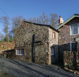 A stone cottage with multiple windows and a gravel driveway surrounded by leafless trees at Church How Cottage in Nether Wasdale
