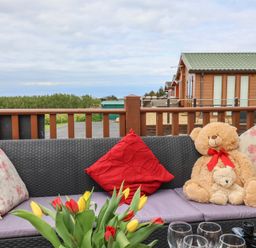 An outdoor seating area with a wicker couch, floral and red cushions, two teddy bears, tulips on a table, glassware, and a green-roofed cabin in the background at Poppy Lodge Mullacott Ilfracombe