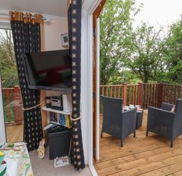 A small living area with a wall-mounted television and shelves beside a deck with two wicker chairs and a small table at Poppy Lodge in Mullacott Ilfracombe
