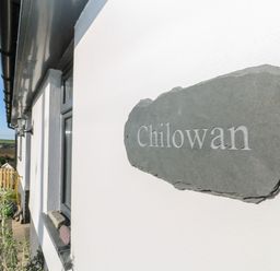 A close up of a stone house name plaque on a white wall with a window and outdoor area at Chilowan in Trewarmett near Tintagel