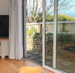 A living room with a television on a stand and sliding glass doors opening to a patio with a stone wall at Chilowan in Trewarmett near Tintagel