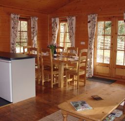 A dining area with wooden chairs and table set with glasses and a bottle next to kitchen counter and coffee table in a wooden room at Valley Lodge Oakfordbridge near Bampton