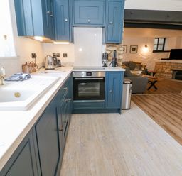 A kitchen with blue cabinets a white countertop a brass faucet and appliances next to a living room with a sofa a TV and a stone fireplace at Ellie's Cottage in Burford