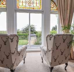 Two armchairs with deer head patterns in front of a large window with stained glass panels and curtains in a sitting room with a plant in a pot at Ismay Dining Room Suite - Titanic Interest in Waterloo Merseyside