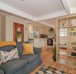 An open plan living area with a blue sofa and yellow cushions a wooden partition shelf and a kitchen with white cabinets at Nuthatch in Honiton