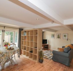A living and dining area with a table and chairs beside a large shelving unit and a sofa with a television on a stand at Nuthatch in Honiton