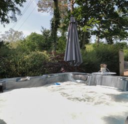 A bubbling hot tub outdoors with a closed umbrella and a small table with a flower pot surrounded by trees and bushes at Nuthatch in Honiton