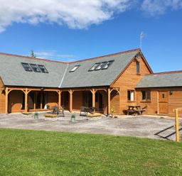 A wooden house with a grey roof and outdoor seating on a paved area with grass in front at Riverside Lodge in Washford
