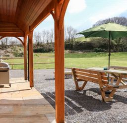 A covered patio with wooden posts a barbecue grill and an outdoor picnic table with a green umbrella in a grassy yard at Riverside Lodge in Washford
