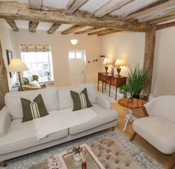 A living room with a sofa, armchair, side table, wooden beams, and a sideboard with lamps at Lower Pack Horse Cottage in Winchcombe