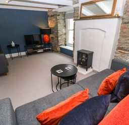 A living room with grey sofas orange and dark blue cushions a black round coffee table a small black stove a television on a stand and a stone wall at Winterbrook Cottage in Mevagissey