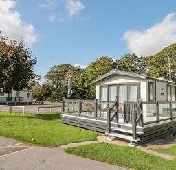 A white static caravan with a gray deck and chairs on a grassy area near a road at The Olive Lodge 227 Sycamore in New Milton